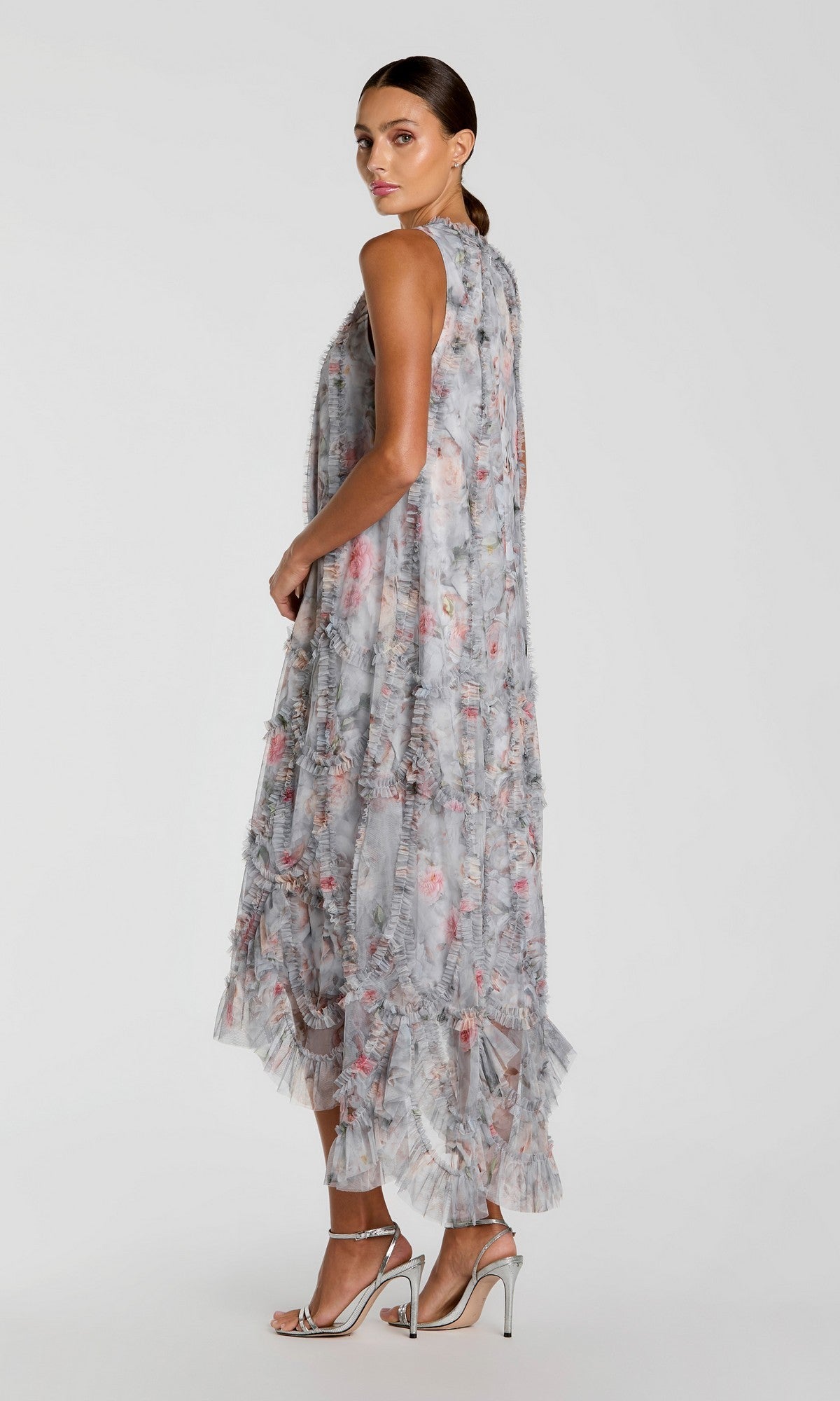A woman in profile models the Mac Duggal 8430 Midi Party Dress—an ankle-length, ruffled floral cocktail dress with sheer layers—paired with silver high-heeled sandals, posed against a plain light background.